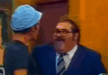 a man with glasses and a blue hat talks to another man in a suit