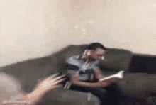 a man is sitting on a couch reading a book while a woman throws a frisbee .