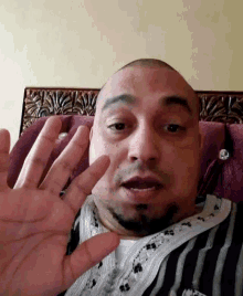 a bald man with a beard is sitting on a couch with his hand up .