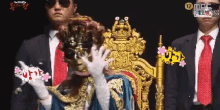 a man in a suit and tie is standing next to a woman in a mask on a throne