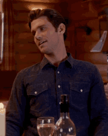 a man in a denim shirt is sitting at a table with two glasses of wine and a bottle of wine .