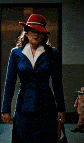 a woman in a blue suit and red hat stands in a hallway