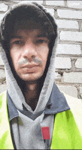 a man wearing a hoodie and a yellow vest stands in front of a white brick wall