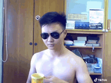 a shirtless man wearing sunglasses holds a cup in front of a wall with a paw print sticker