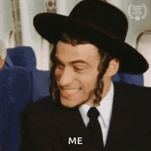 a man wearing a hat and tie is smiling and saying `` me '' while sitting on an airplane .