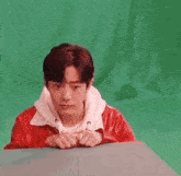 a young man in a red jacket is sitting in front of a green screen and making a funny face .