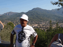 a man wearing a white hat and a black and white shirt with a dragon on it stands in front of a mountain range