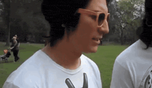 a man wearing sunglasses stands in a park with a stroller in the background