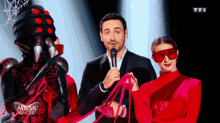 a man in a suit holds a microphone in front of a masked singer