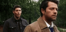 two men are standing next to each other in a forest . one of the men is wearing a suit and tie .