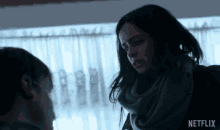 a woman in a scarf talks to a man in front of a window with a netflix logo in the corner