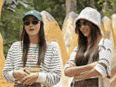 a woman wearing a la hat stands next to another woman wearing sunglasses and a striped shirt