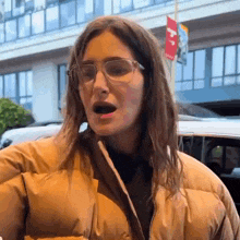 a woman wearing glasses and a puffy jacket is standing in front of a building .