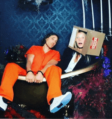 a man in a jail uniform sits next to a woman in a box that says fragile on it