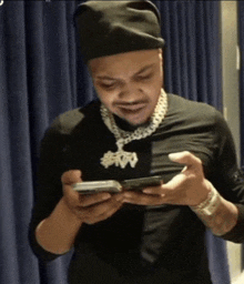 a man wearing a black hat and a necklace that says $ 100 looks at his phone