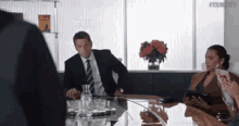 a man in a suit and tie is sitting at a table with a woman sitting behind him .