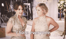 two women in gold dresses are standing next to each other and one of them says " i hate everyone "