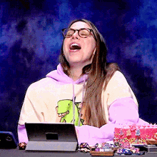 a woman wearing glasses and a sweatshirt with a dinosaur on it is laughing in front of a tablet