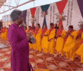 a man in a purple robe is standing in front of a group of people in yellow dresses