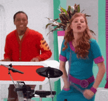 a woman in a blue dress is playing a drum set while a man in a red jacket stands behind her .