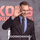 a man in a suit and tie is waving at the camera while standing on a red carpet .