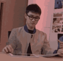 a young man wearing glasses is sitting at a table with a tablet .