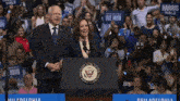 a man and woman stand at a podium in front of a crowd with harris walz signs