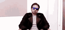 a man wearing sunglasses and a leather jacket is sitting in a chair making a face .