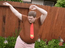 a man wearing a brown shirt and a red tie with the letter b on it is dancing in front of a wooden fence