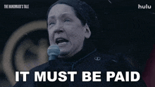 a woman speaking into a microphone with the words it must be paid behind her