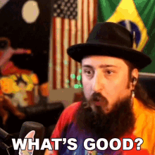 a man with a beard wearing a hat says " what 's good " in front of a microphone