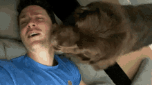 a man in a blue shirt is laying on a bed with a brown dog licking his face