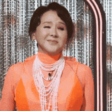a woman in an orange dress and pearls is standing in front of a neon sign .