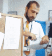a man with a beard is standing in front of a bulletin board looking at a piece of paper .