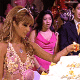 a woman in a pink dress is cutting a cake with a peace sign on her necklace