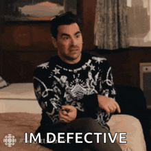 a man in a sweater is sitting on a bed and says im defective .