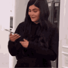 a woman in a black sweater is standing in front of a refrigerator and looking at her phone .