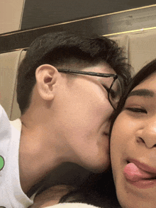 a man with glasses kisses a woman on the cheek while she sticks her tongue out