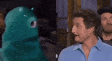 a man in a blue shirt is standing next to a green stuffed animal and talking to it .