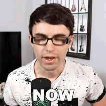 a man wearing glasses is talking into a microphone with the word now written on it
