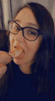 a woman wearing glasses is eating a piece of food with her tongue out