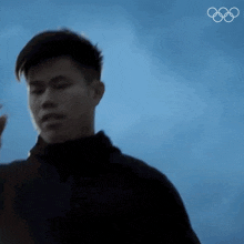 a man in a black jacket is standing in front of a blue sky with the olympic rings in the background