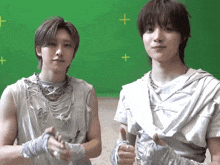 two young men are standing in front of a green screen with a plus sign