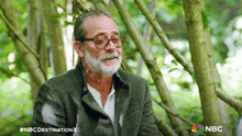 a man with glasses and a beard is sitting in a forest with a nbc logo in the corner