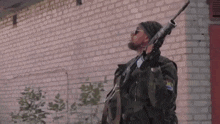 a man with a beard is holding a gun in front of a brick building .