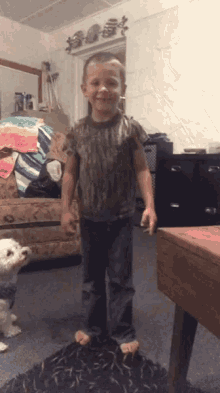 a young boy in a camouflage shirt stands in a living room next to a dog