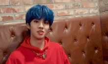 a young man with blue hair and a red hoodie is sitting on a leather couch .