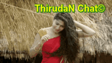 a woman in a red saree is standing in front of a thatched roof and looking at her hair .