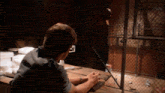 a man sits at a table in front of a chain link fence looking out a window
