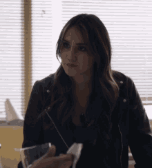 a woman wearing a black leather jacket is sitting in front of a window with blinds